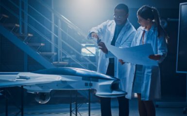 Two Aerospace Engineers Work On Unmanned Aerial Vehicle / Drone Prototype. Aviation Scientists in White Coats Holding Blueprints. Laboratory with Commercial Aerial Surveillance Aircraft